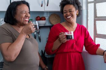 Two Black women laughing in a kitchen with cups of black coffee in hand.
