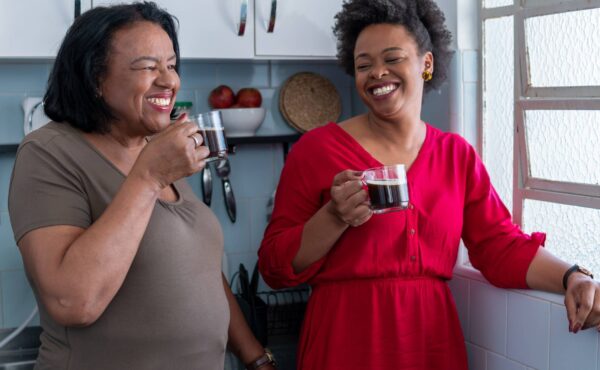 Two Black women laughing in a kitchen with cups of black coffee in hand.