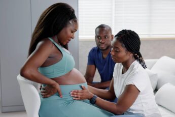 A doula feeling the stomach of a pregnant woman who is sitting on a chair with her husband in the background.