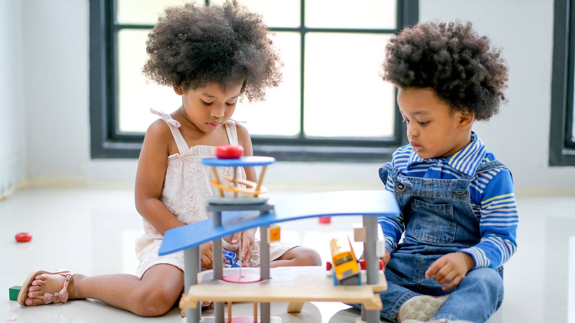 A little girl and her brother sit on the floor in a white room and play together with a toy building, bus, and blocks.