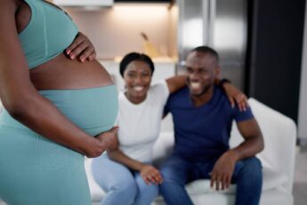 A pregnant black surrogate mother holding her belly as she stands in front of a smiling black couple sat on a white couch.