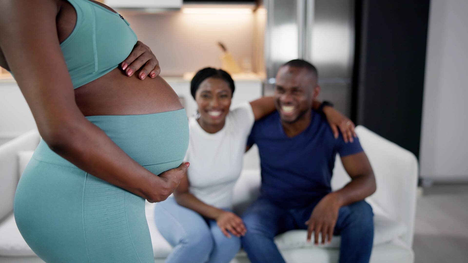 A pregnant black surrogate mother holding her belly as she stands in front of a smiling black couple sat on a white couch.