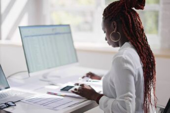 A Black woman sitting at a desk with dual-screens, reviewing tax documents to get herself set up for next filing season.