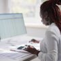 A Black woman sitting at a desk with dual-screens, reviewing tax documents to get herself set up for next filing season.