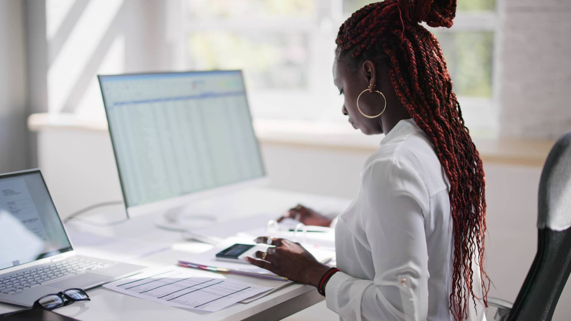 A Black woman sitting at a desk with dual-screens, reviewing tax documents to get herself set up for next filing season.