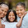 A smiling, young Black girl standing between her grandmother and mother. She is cupping their cheeks as they lean in.