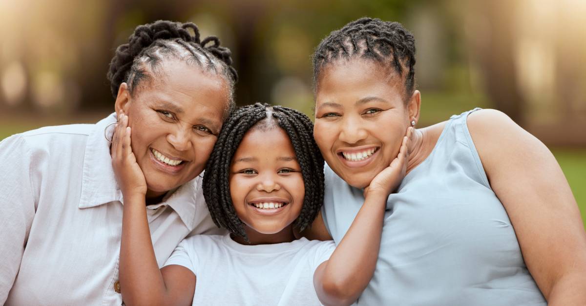 A smiling, young Black girl standing between her grandmother and mother. She is cupping their cheeks as they lean in.