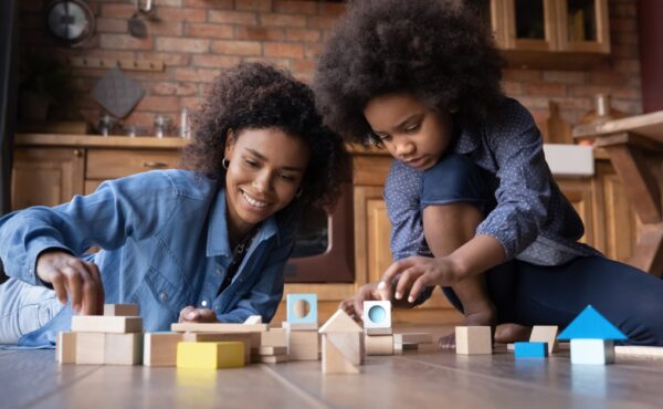 A mother and her young daughter are sitting together on the floor in a kitchen and playing with wooden blocks.