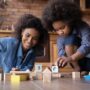 A mother and her young daughter are sitting together on the floor in a kitchen and playing with wooden blocks.