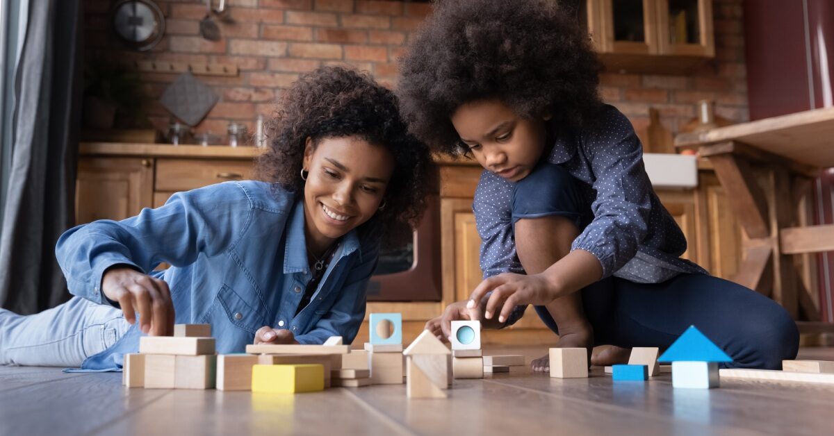 A mother and her young daughter are sitting together on the floor in a kitchen and playing with wooden blocks.