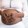 A Black woman sitting on a bed with her shirt lifted to show her pregnant belly; her hands are on her tummy in a heart shape.