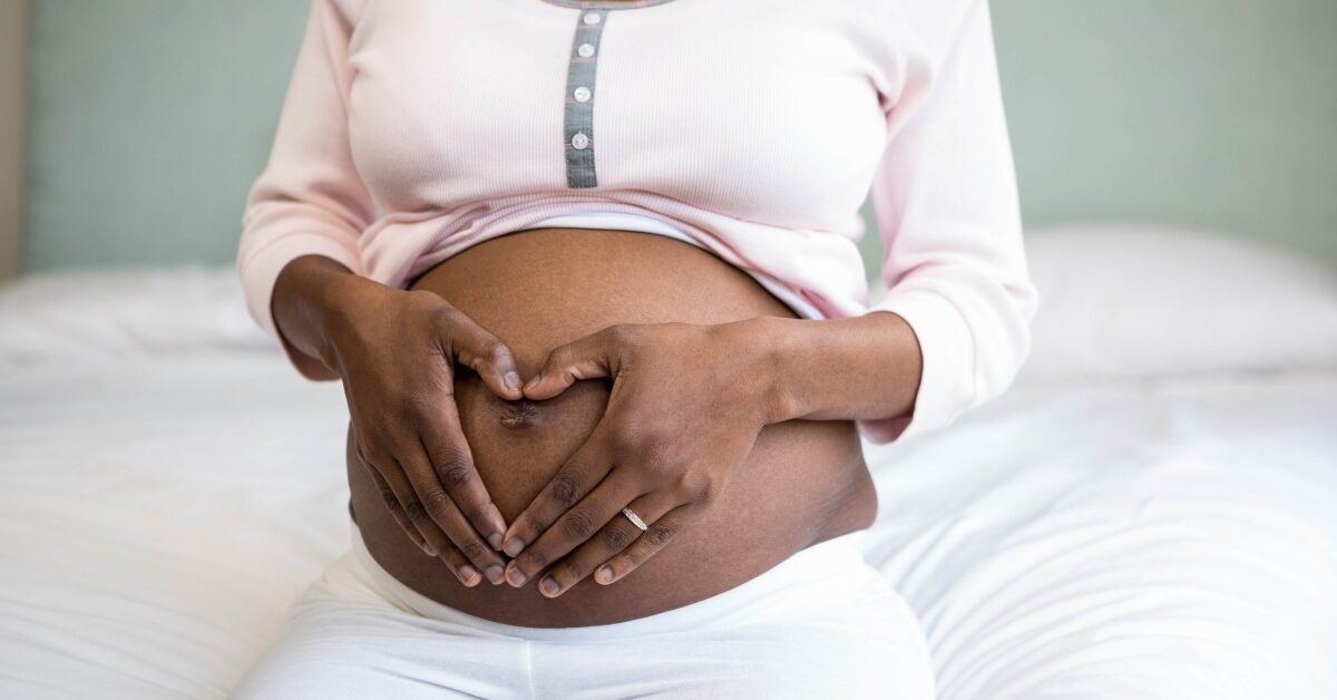 A Black woman sitting on a bed with her shirt lifted to show her pregnant belly; her hands are on her tummy in a heart shape.