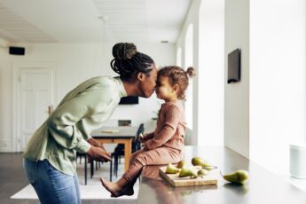 A mother and a daughter are rubbing noses and smiling at each other. The daughter is sitting on the counter eating a snack.