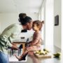 A mother and a daughter are rubbing noses and smiling at each other. The daughter is sitting on the counter eating a snack.