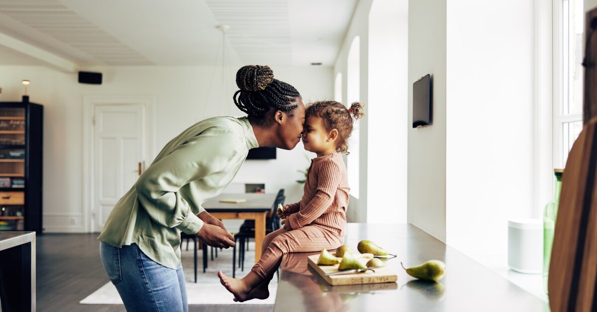 A mother and a daughter are rubbing noses and smiling at each other. The daughter is sitting on the counter eating a snack.