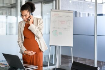 A pregnant woman holding her tummy as she talks on the phone and looks at her computer in her corporate office.