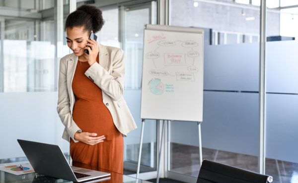 A pregnant woman holding her tummy as she talks on the phone and looks at her computer in her corporate office.