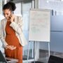 A pregnant woman holding her tummy as she talks on the phone and looks at her computer in her corporate office.