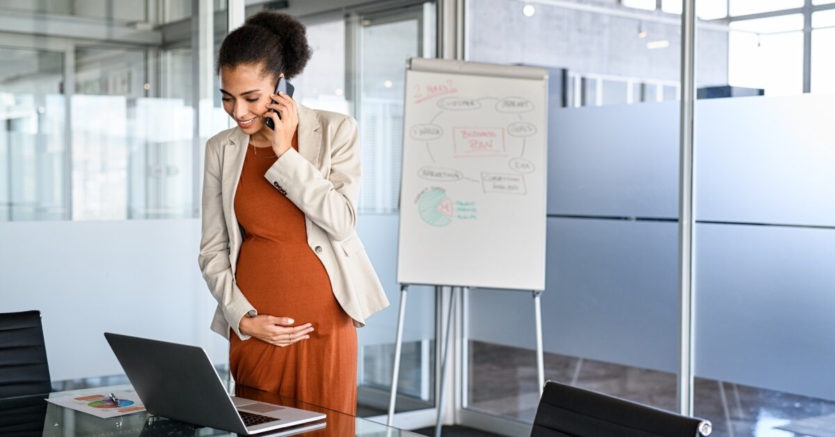 A pregnant woman holding her tummy as she talks on the phone and looks at her computer in her corporate office.