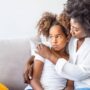 A concerned mother hugging her daughter on a couch. The daughter looks worried, holding her hands in her lap.