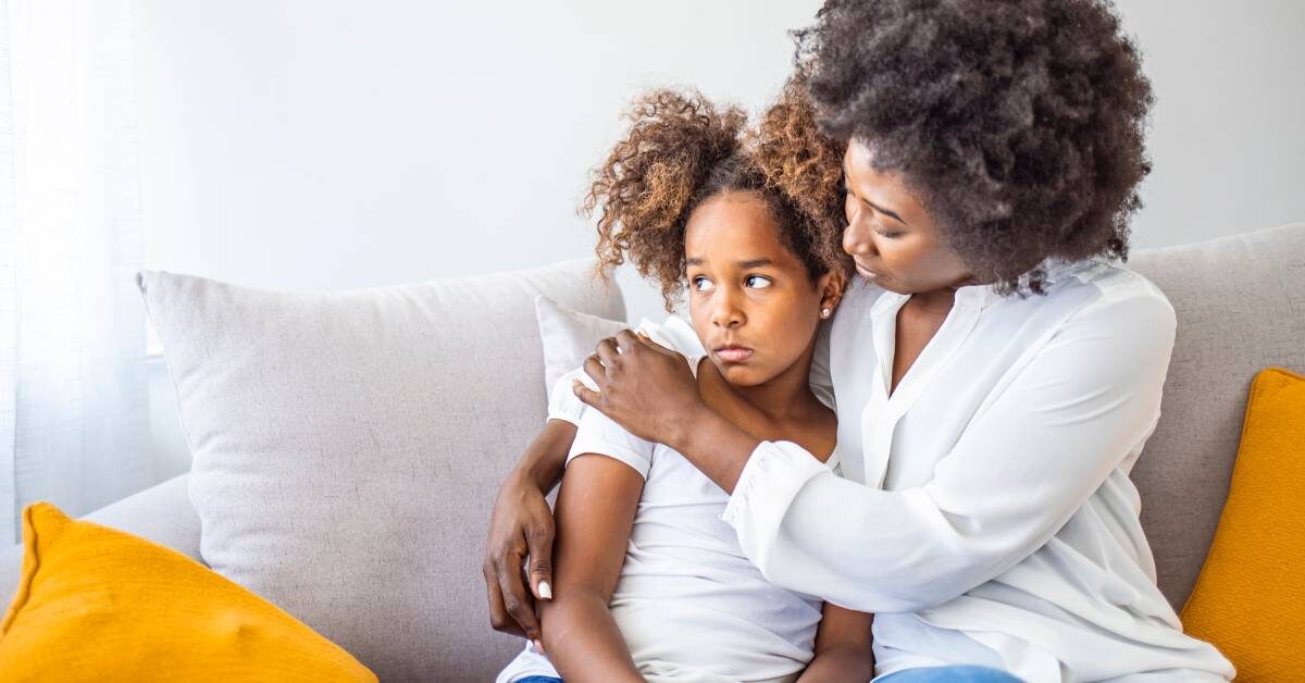 A concerned mother hugging her daughter on a couch. The daughter looks worried, holding her hands in her lap.