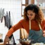 A woman reaching for an ingredient on the counter with a propped-up tablet, a cutting board, a mixing bowl, and ingredients.