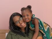 A Black mother sitting on a bench and smiling at the camera while her cute daughter leans over her shoulder.