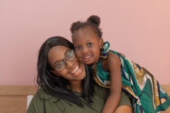 A Black mother sitting on a bench and smiling at the camera while her cute daughter leans over her shoulder.