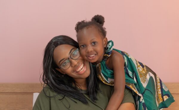 A Black mother sitting on a bench and smiling at the camera while her cute daughter leans over her shoulder.