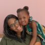 A Black mother sitting on a bench and smiling at the camera while her cute daughter leans over her shoulder.