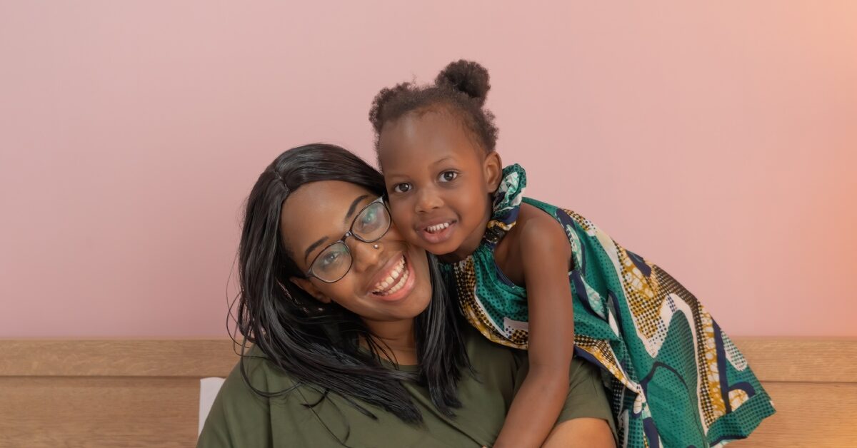 A Black mother sitting on a bench and smiling at the camera while her cute daughter leans over her shoulder.