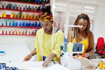 A Black mom and daughter duo working closely on a sewing craft. There's a shelf with tons of spools hanging behind them.