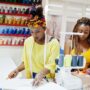 A Black mom and daughter duo working closely on a sewing craft. There's a shelf with tons of spools hanging behind them.