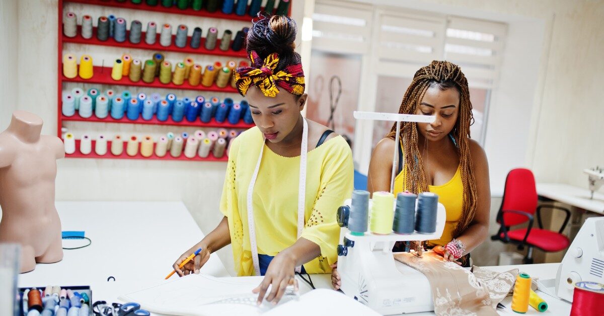 A Black mom and daughter duo working closely on a sewing craft. There's a shelf with tons of spools hanging behind them.