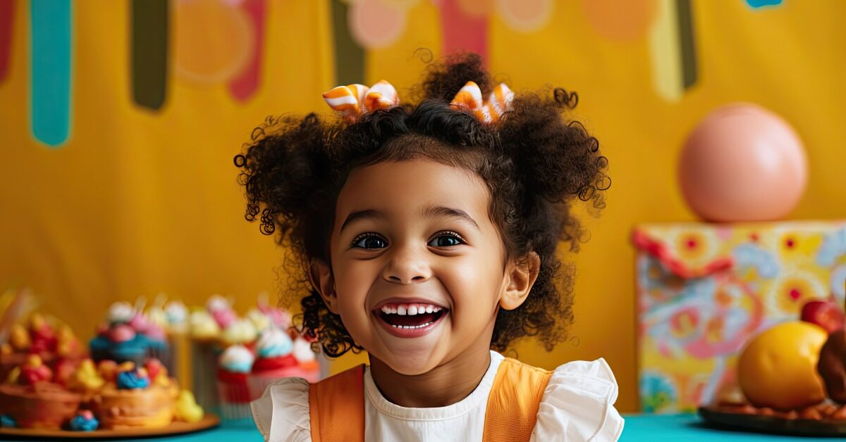 A little girl at a birthday party has a large grin. She's wearing orange and white striped bows in her hair.