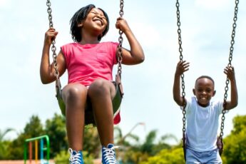 Two joyful Black children swinging on swings in a vibrant park surrounded by green trees and playground equipment.