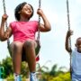 Two joyful Black children swinging on swings in a vibrant park surrounded by green trees and playground equipment.