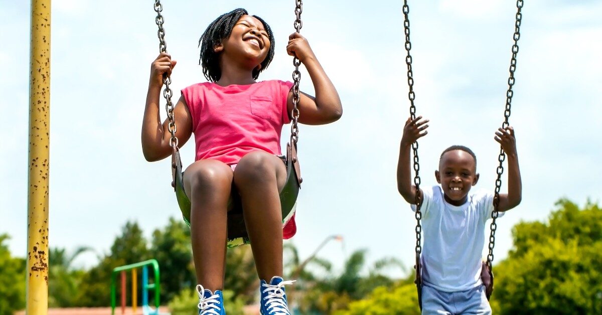 Two joyful Black children swinging on swings in a vibrant park surrounded by green trees and playground equipment.