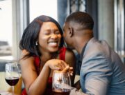 An African American couple smiling, holding hands, and enjoying glasses of wine at a romantic date night in a restaurant.