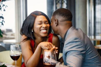 An African American couple smiling, holding hands, and enjoying glasses of wine at a romantic date night in a restaurant.