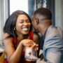 An African American couple smiling, holding hands, and enjoying glasses of wine at a romantic date night in a restaurant.