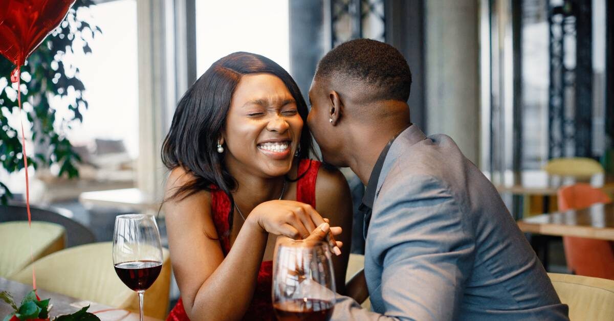 An African American couple smiling, holding hands, and enjoying glasses of wine at a romantic date night in a restaurant.