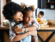 A black mother and her daughter are smiling and laughing in the kitchen while looking off to the side.