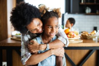 A black mother and her daughter are smiling and laughing in the kitchen while looking off to the side.