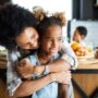 A black mother and her daughter are smiling and laughing in the kitchen while looking off to the side.