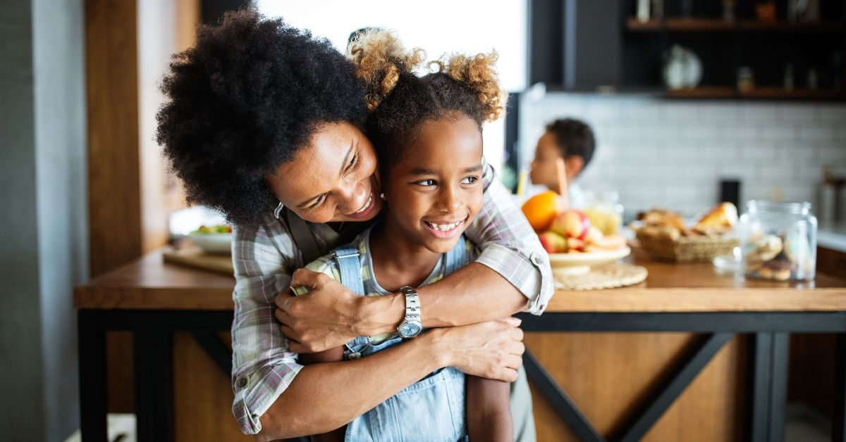 A black mother and her daughter are smiling and laughing in the kitchen while looking off to the side.