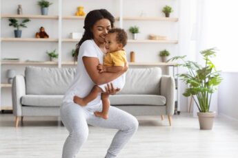 A mother is standing on an exercise mat and performing a squat. She is holding her smiling baby in her arms.