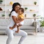 A mother is standing on an exercise mat and performing a squat. She is holding her smiling baby in her arms.