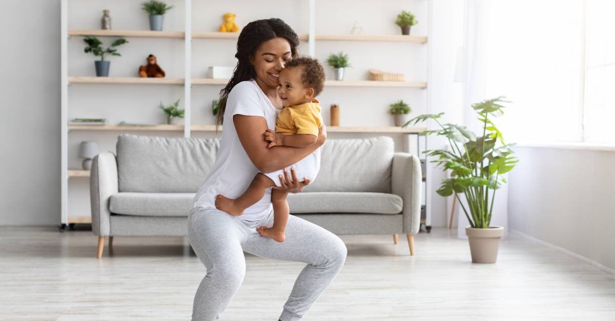 A mother is standing on an exercise mat and performing a squat. She is holding her smiling baby in her arms.