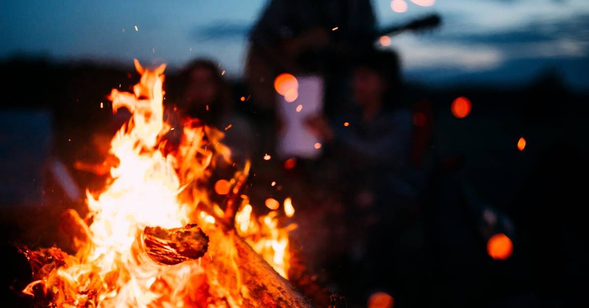 A small bonfire, crackling with red-hot flames. A family sits and stands around the fire. It's late at night.